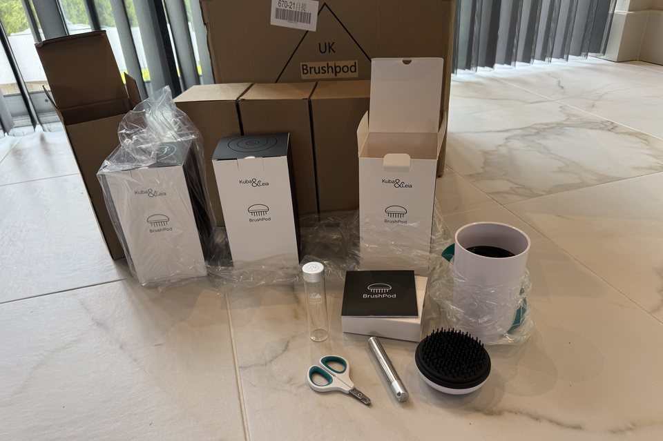 Three BrushPod retail cartons standing in front of stacked trade boxes, with one unit unpacked alongside to show the brush, claw clippers, laser toy, presentation canister and glass tube